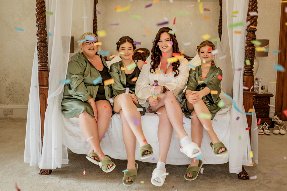 Bride and her bridesmaids do confetti shot sitting on four poster bed